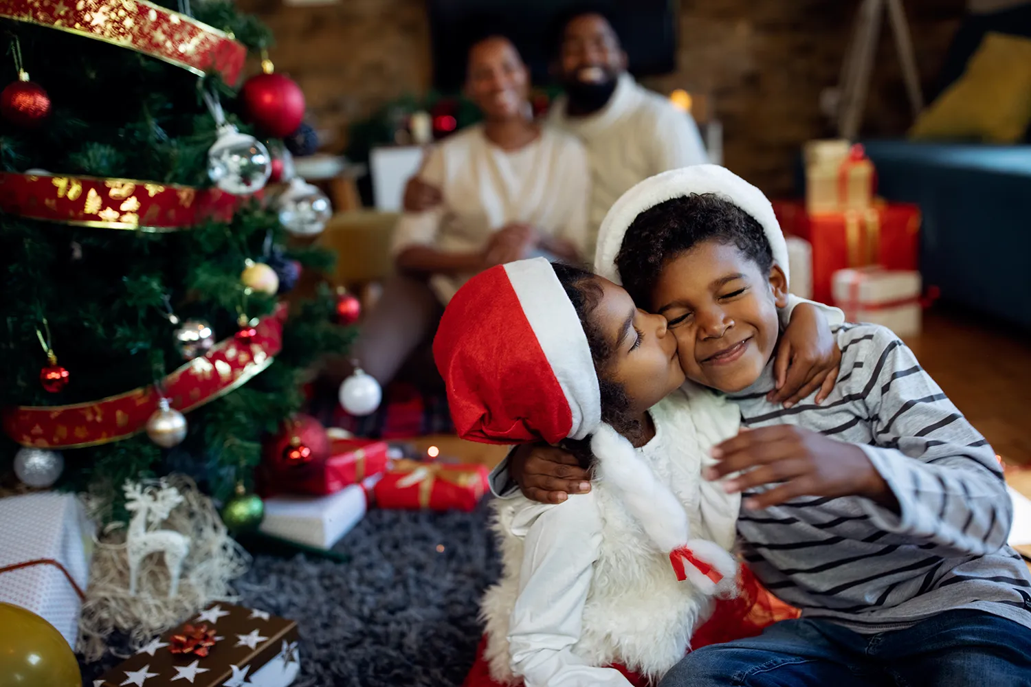 Irmã e irmão de abraçando ao lado de arvore de Natal, com os pais no fundo sorrindo
