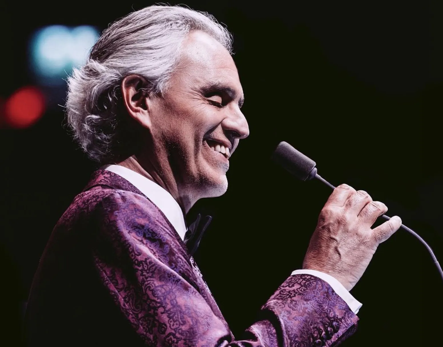 Andrea Bocelli - Brasil 2024 - Dançar Marketing - Just Real Moms Close de Andrea Bocelli no palco sorrindo com microfone na mão usando blaser lilás