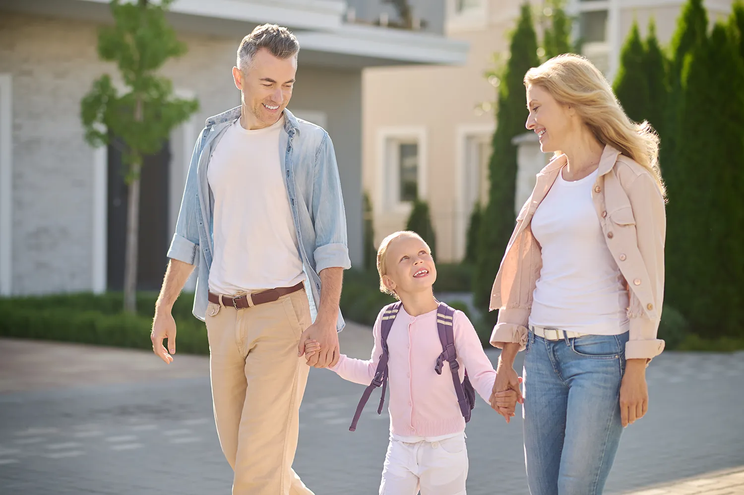 Pais e filha - Volta às aulas 2024 - Lista de materiais - Just Real Moms Mãe e pai de mãos dadas com filha pequena levando-a para a escola na volta as aulas