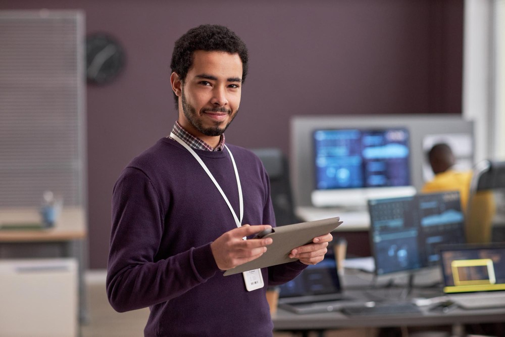Gestor de automação sorrindo em ambiente de trabalho com computadores e gráficos ao fundo.